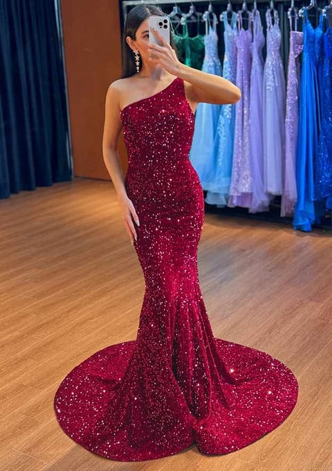 Woman in burgundy sequined one-shoulder mermaid gown taking a mirror selfie in a dressing room with colorful formal dresses displayed in background